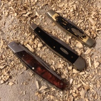 A set of three slightly different closed whittling knives, laying in a row diagonally on a beige canvas tarp, surrounded by wood shavings.