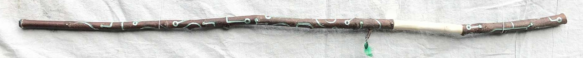 A horozontal photo of the staff, dark brown bark, peeled grip, and a green feather hanging from an accessory branch, with a canvas backdrop. Shimmering green circuitboard like linework is carved along the length of it.