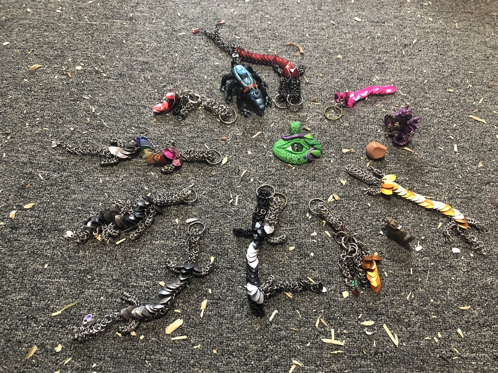 Thirteen dragons surrounding a green dragon eye pendant on a wood chip covered grey carpet, all looking at Paul, the newest adopted Dragon.