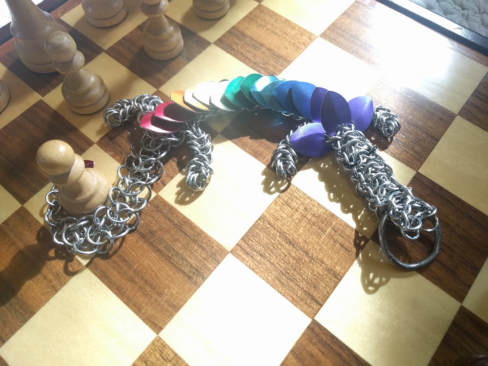 A purple, blue, light blue, green, yellow, orange, and red striped dragon laying atop a chess board, with the first white pawn moved up, held in her tail.