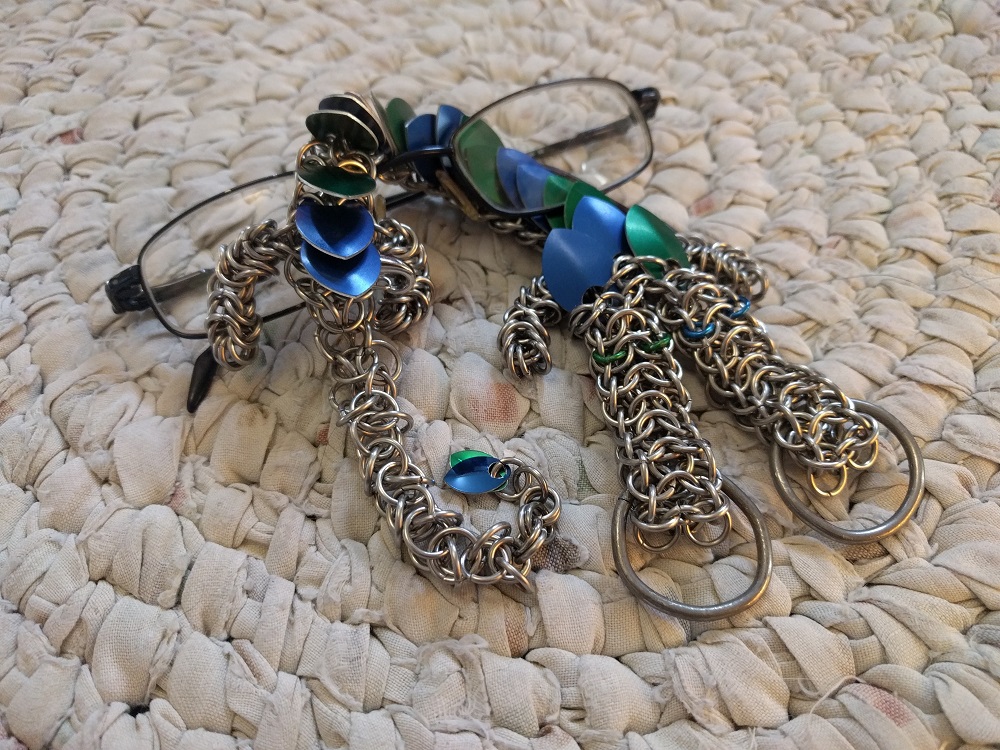 A two headed blue and green striped dragon, wrapped around a pair of glasses and sitting on a white crocheted rug.