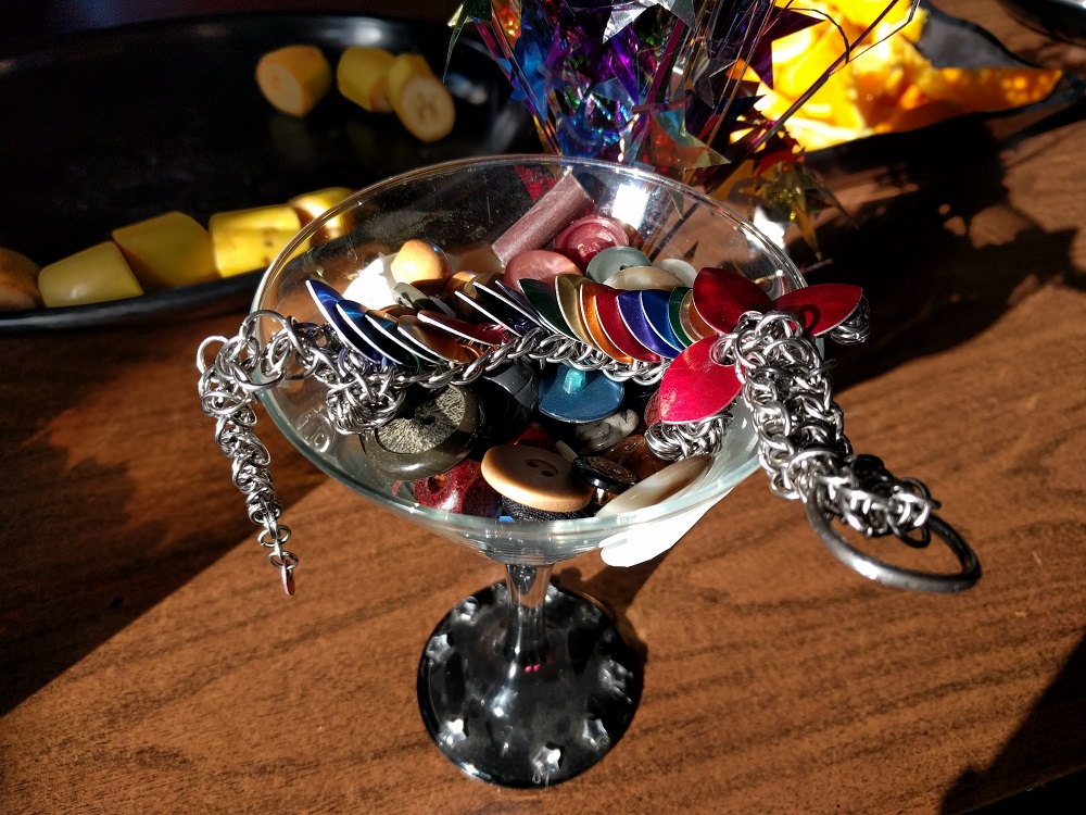 A triple rainbow striped dragon sitting inside of a martini glass that is half filled with buttons, on top of a coffee table with candies and a celebration ornament behind her.