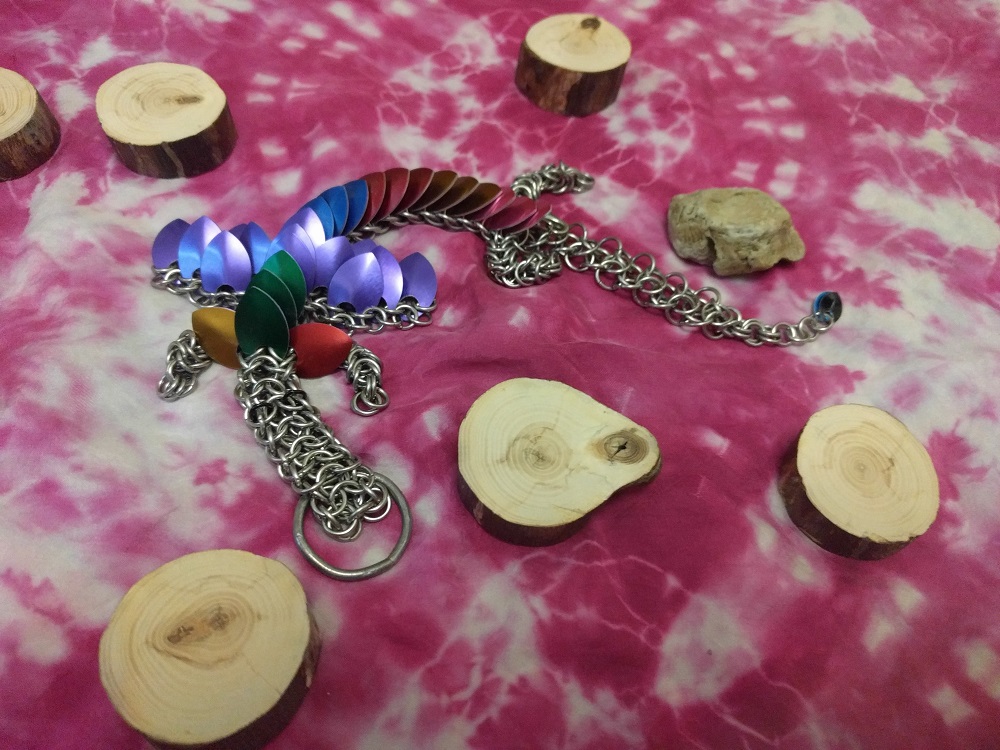 A green, purple, blue, red, brown, and pink striped dragon with brown and red neck frills and purple wings laying atop a pink divination fabric surrounded by the wooden and stone pieces of the divination kit.