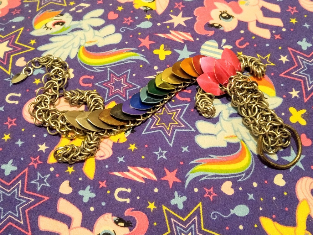 A pink, red, orange, brown, yellow, light green, green, light blue, blue, purple, black, rusty, dark grey, silver, titanium striped dragon, showing all of the colours I have had available, dragon, sitting on top of a purple, very busy with stars and horseshoes and dots my little pony pillowcase.