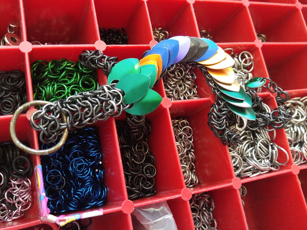 A green, orange, blue, purple, black, purple, orange, yellow, green striped dragon laying on top of my open chainmaille kit.