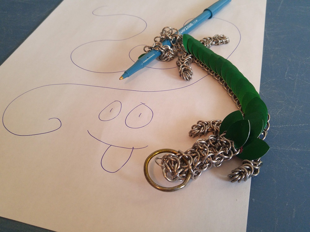 A green dragon holding a blue pen in his coiled up tail, laying on top of a paper with random squiggles on it, and a smiling face with its tongue sticking out.