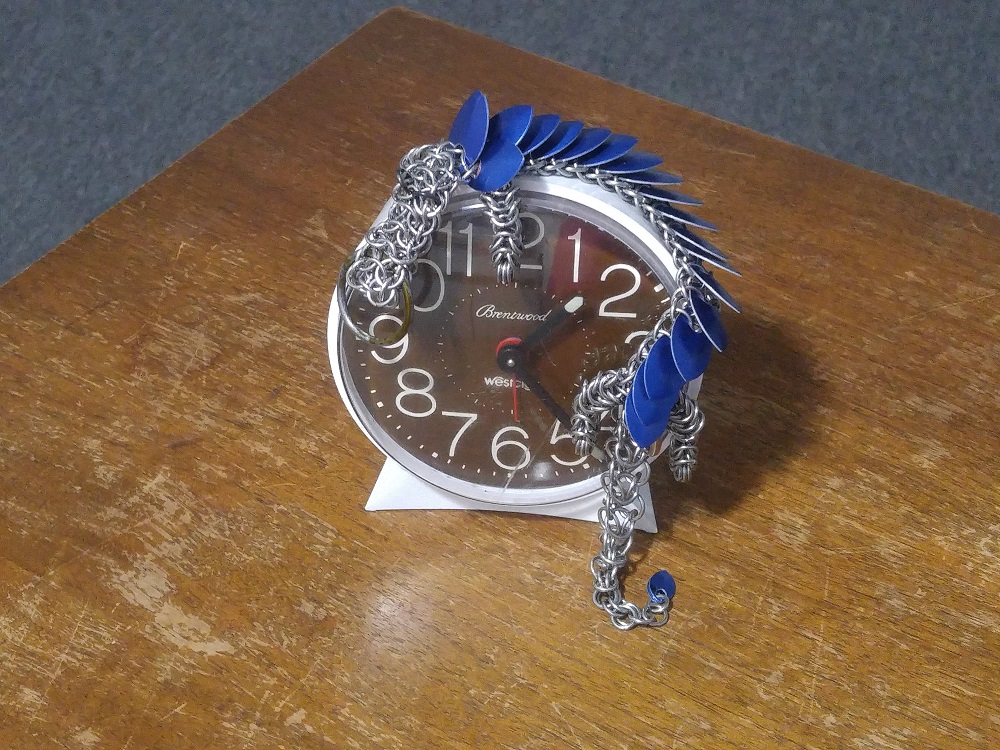 A blue dragon curled on top of a white alarm clock, sitting on a wooden table.