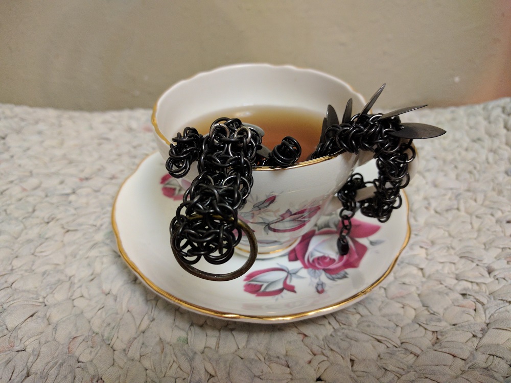 A black dragon with black scales literally sitting inside of a cup of tea, his head and tail out, waving at the camera.