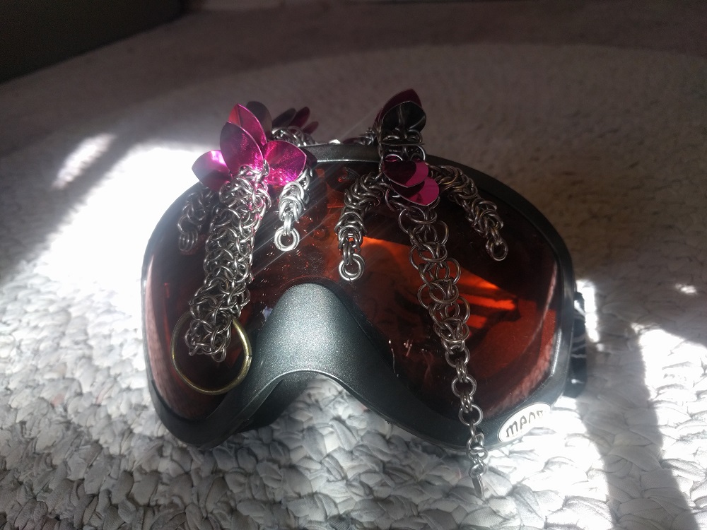 A bright pink and silver striped dragon laying on top of a pair of ski goggles in the sun.