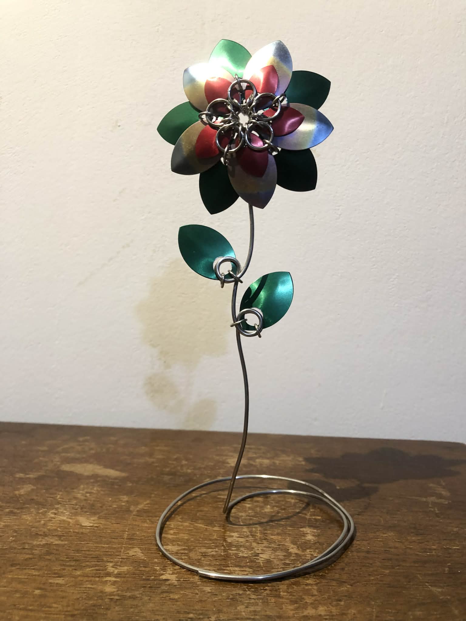 A chainmaille flower, made up of five large rainbow scales with small red scales over them, with large green scales behind, at the end of a long wire stem with two green scale leaves on it.