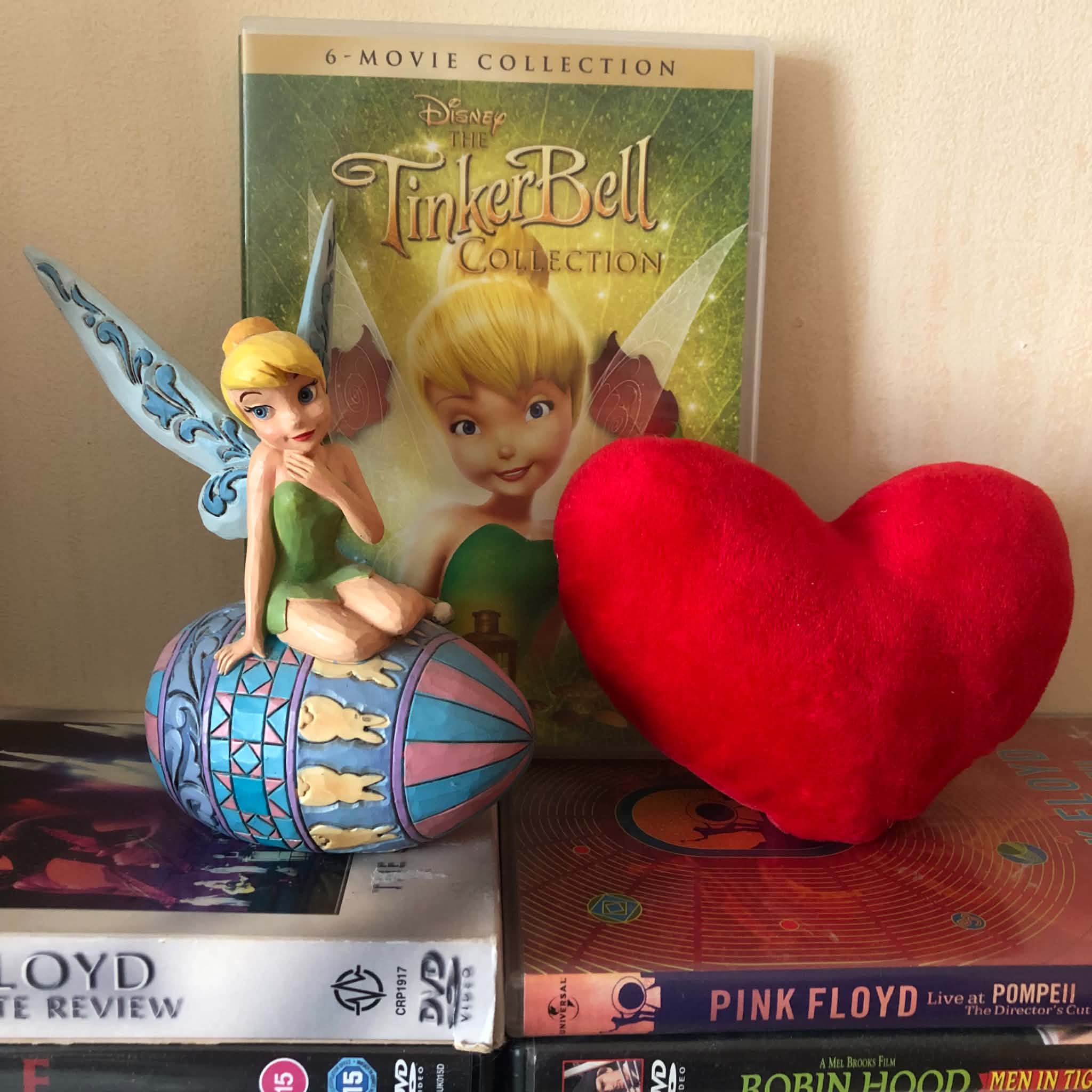 A Tinkerbell figure, movie collection, and a plush heart sitting on top of Pink Floyd dvds.