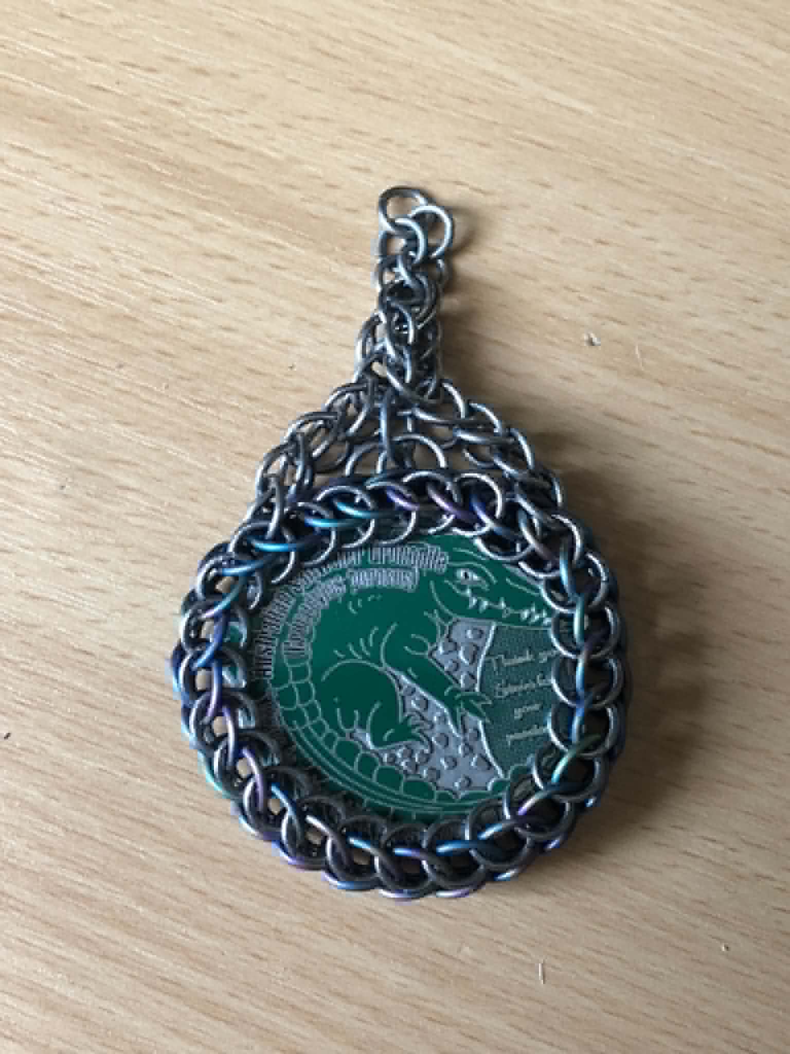 The chainmaille wrapped coin on the workbench, with the half persian strands attached to the wrap, merging into a full persian. Between the full persian is a set of three rings, the top one attached at the base where the half persians connect, and the two lower rings in amongst the coin wrap to tighten it up, and all three of those connected together in a spread out mobius ring style connection.