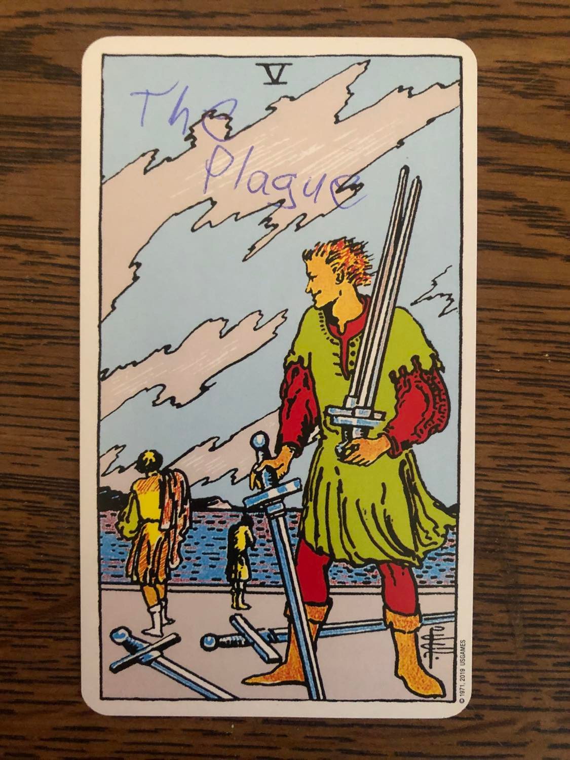 The five of swords card with the plague written on it, laying on a wooden desk.