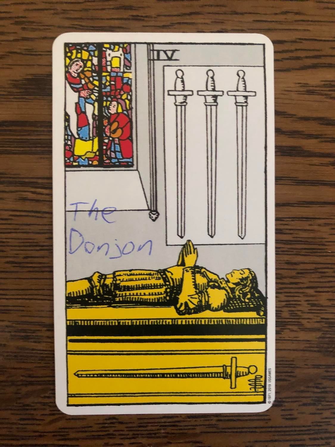 The four of swords card with the donjon written on it, laying on a wooden desk.