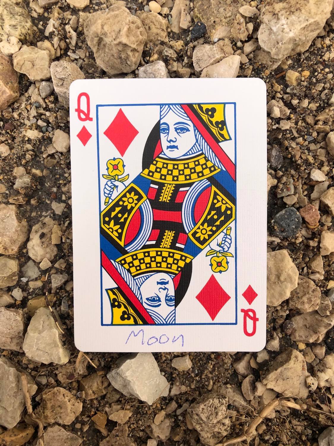 The queen of diamonds with Moon written on it, laying on the gravel path.