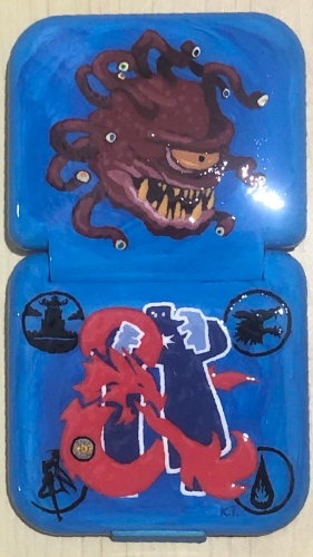 The unfolded light blue box sitting on a wooden table, the top half painted with a red beholder with large spikey teeth looking to the right, its ten eye stalks mainly looking in the same direction, and the bottom half painted with a red Dungeons and Dragons ampersand and blue Paizo golem logo mashup of the two intertwined. The ampersand has a HeroQuest H Q tattoo on the bottom left curve, and the four corners of the back have logo symbols in black. The top left has the Legend of the Five Rings Hiruma clan mon, the top right is the Vampire the Masquerade Gangrel clan symbol, the bottom left has a Sailor Moon silhouette inside the crescent moon, and the bottom right has the Dungeons and Dragons sorcerer symbol.