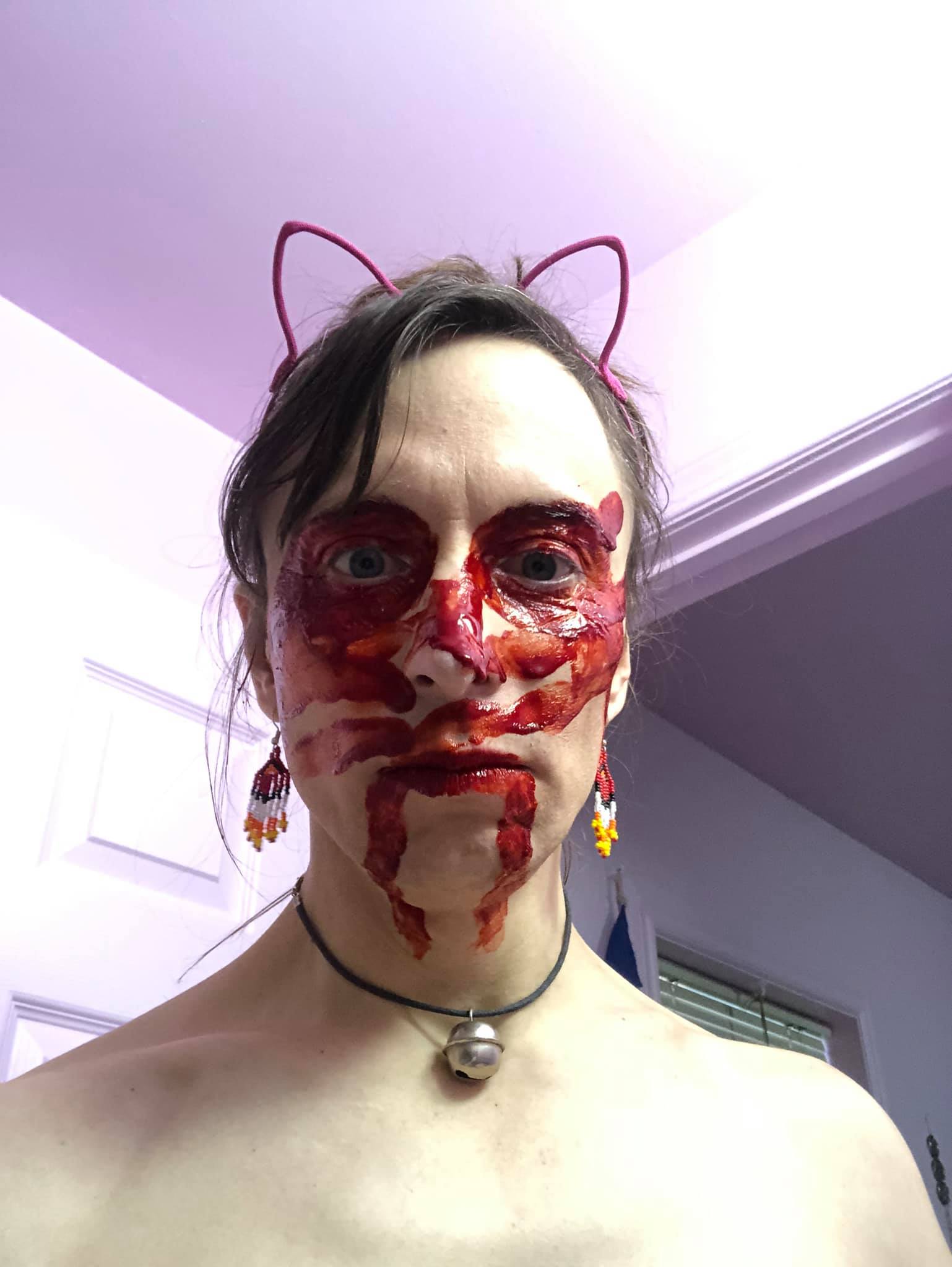 The author, Kabutroid, or Kabuto right now. She has cat ears on, a bell is around her neck, she has red indigenous earrings on, and her face has been painted with blood, running from the corners of her mouth down her throat, her eyes have been painted red, and she has three streaks going down across her face. She seems undisturbed.