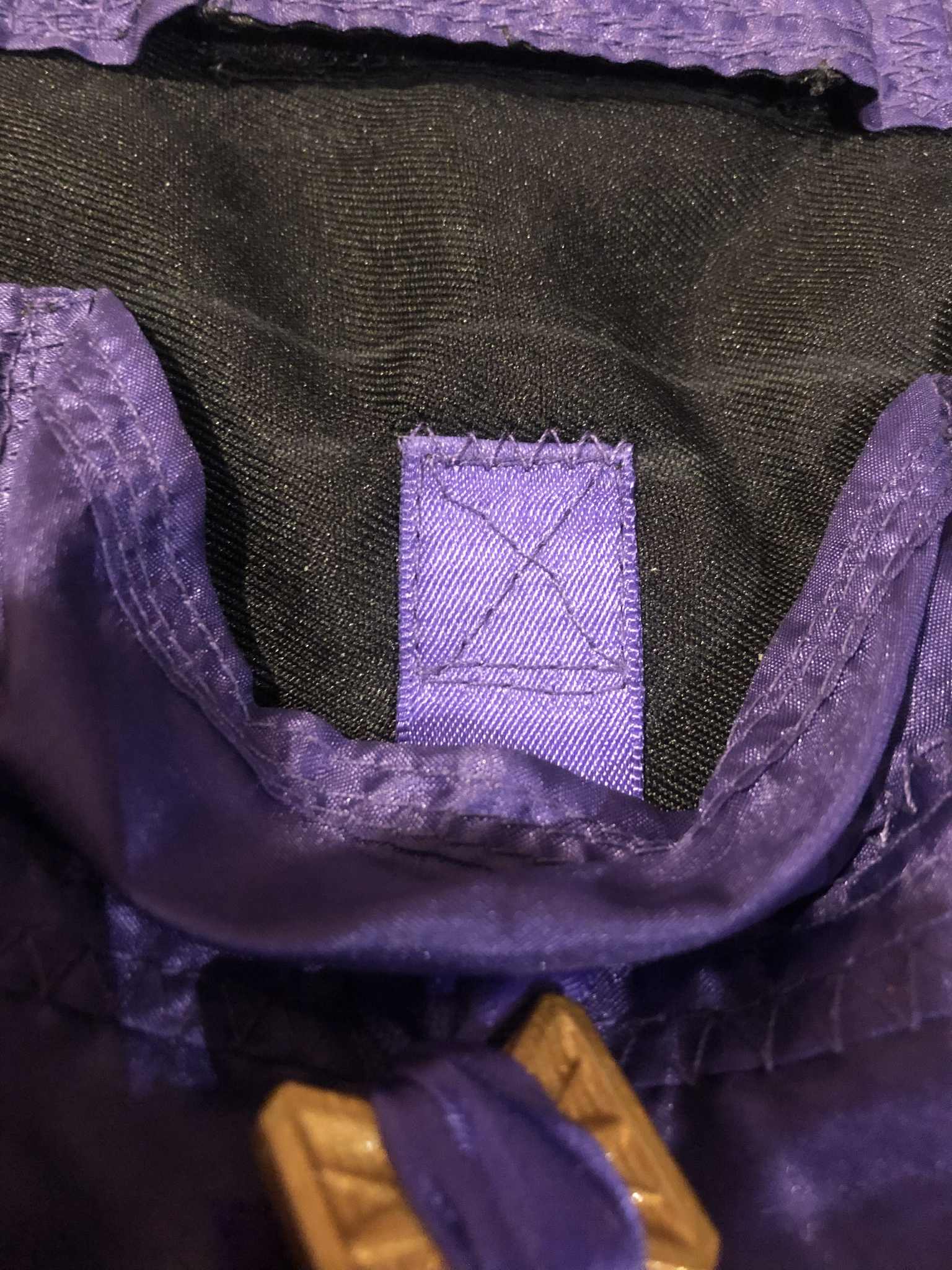 A view of the ribbon attached to the back inside of the pocket, with the chalk lines still visible marking the top of the pocket, and the top of the ribbon a centimeter below that, with a square stitched with an x going through the middle, and the tops and bottoms of the square doubled over, and a zig-zag stitch over the top of the end of the ribbon for smooth sliding. A gold coin is tied to the end of this ribbon.