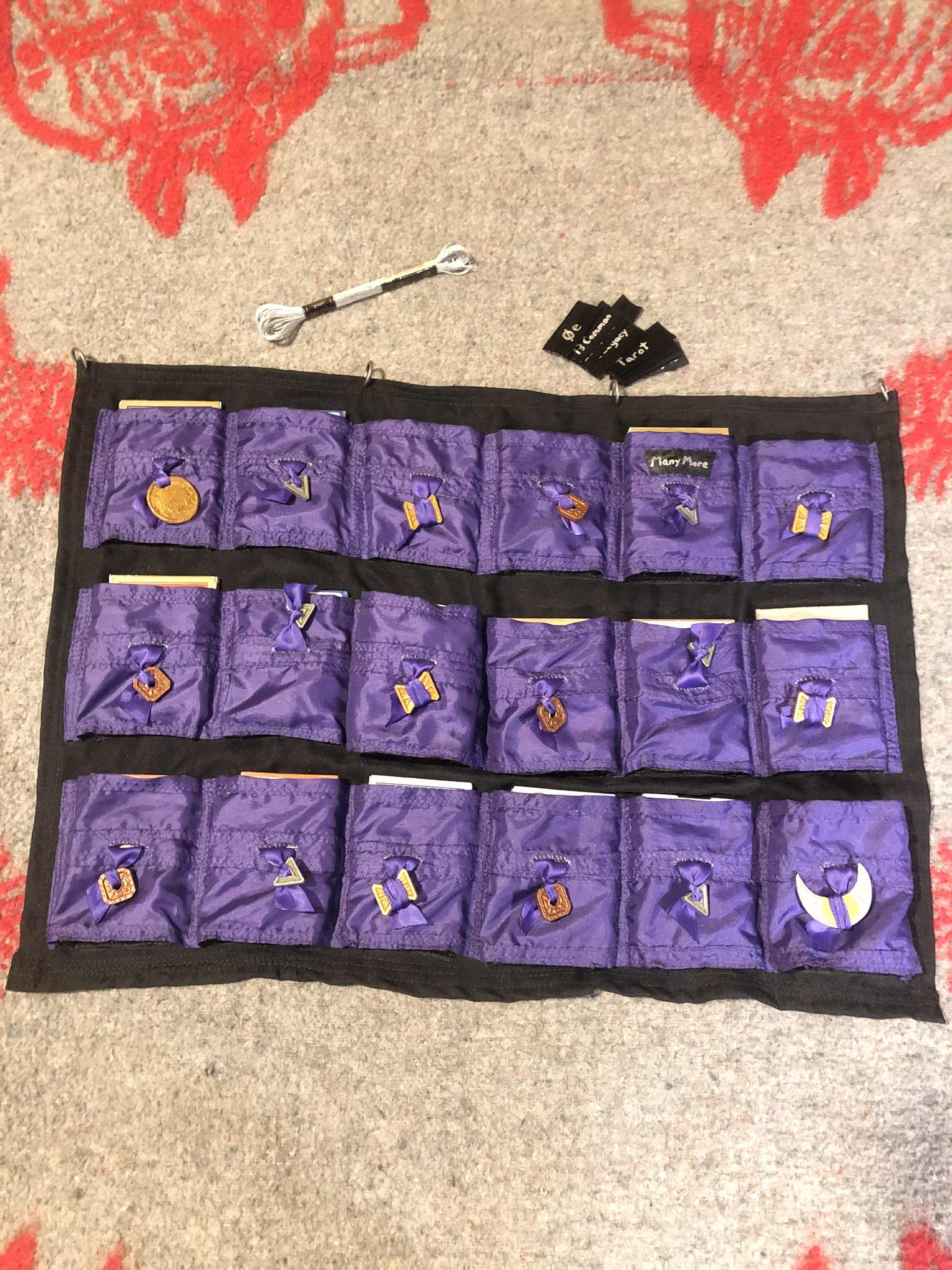 A photo of the full deck sleeve project, with all coins and ribbons attached, and the one label saying Many More attached to the top of the pocket for the deck of many more things. Above the cardsleeve is the remainder of the labels, and the spool of silver embroidery thread that will be used to make the asterisk stars here and there.