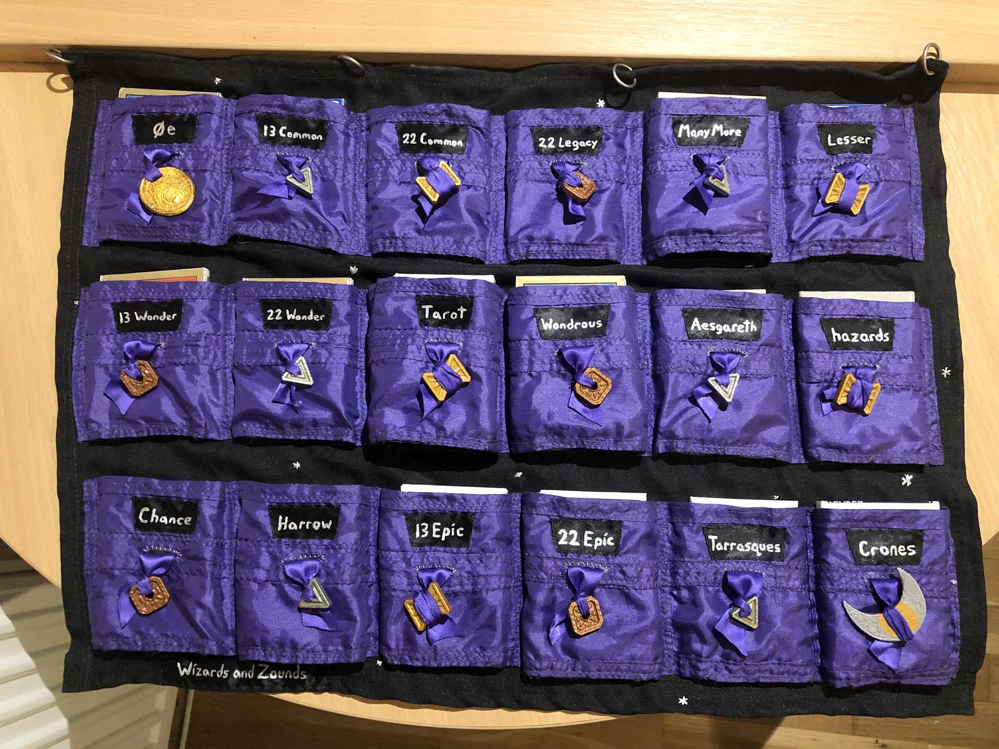 The cardsleeve, fully laid out on a table, each of the pockets filled with their respective decks, with the exception of the very last pocket as only seventeen decks have been discovered, three of them being homebrew. Silver sparkles dot the fabric, labelled as belonging to Wizards and Zounds on the bottom left.