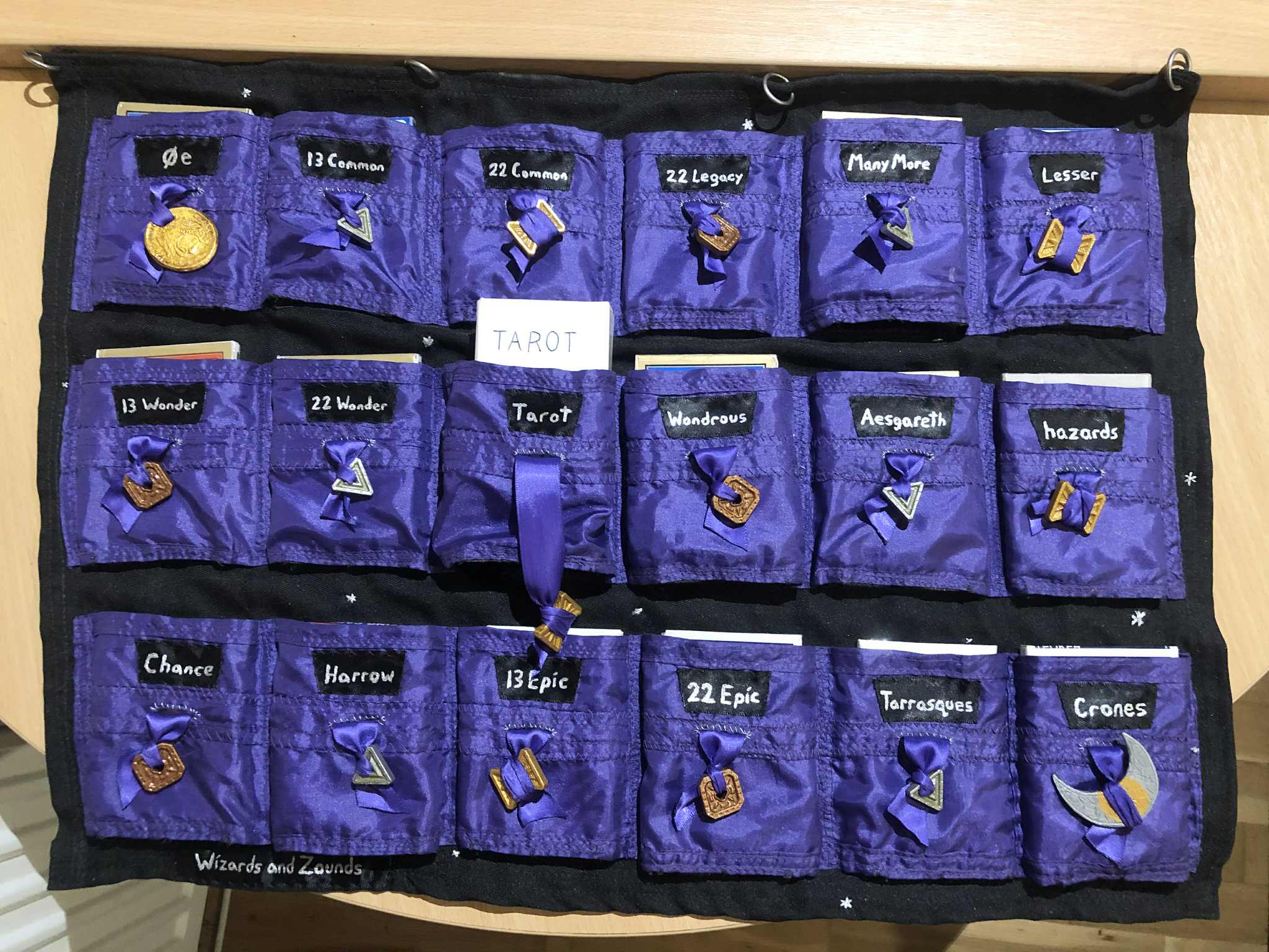 A fabric sheet with many pockets, eighteen total in three rows, though the last row has no label on the last pocket. Shimmery purple fabric on black, each with a drawstring tipped with a dungeons and dragons coin, and the pockets labelled things like zero e, 13 common, 22 wonder, wondrous, harrow, 22 epic, and more. All of the random draw type decks of many things from Dungeons and Dragons.