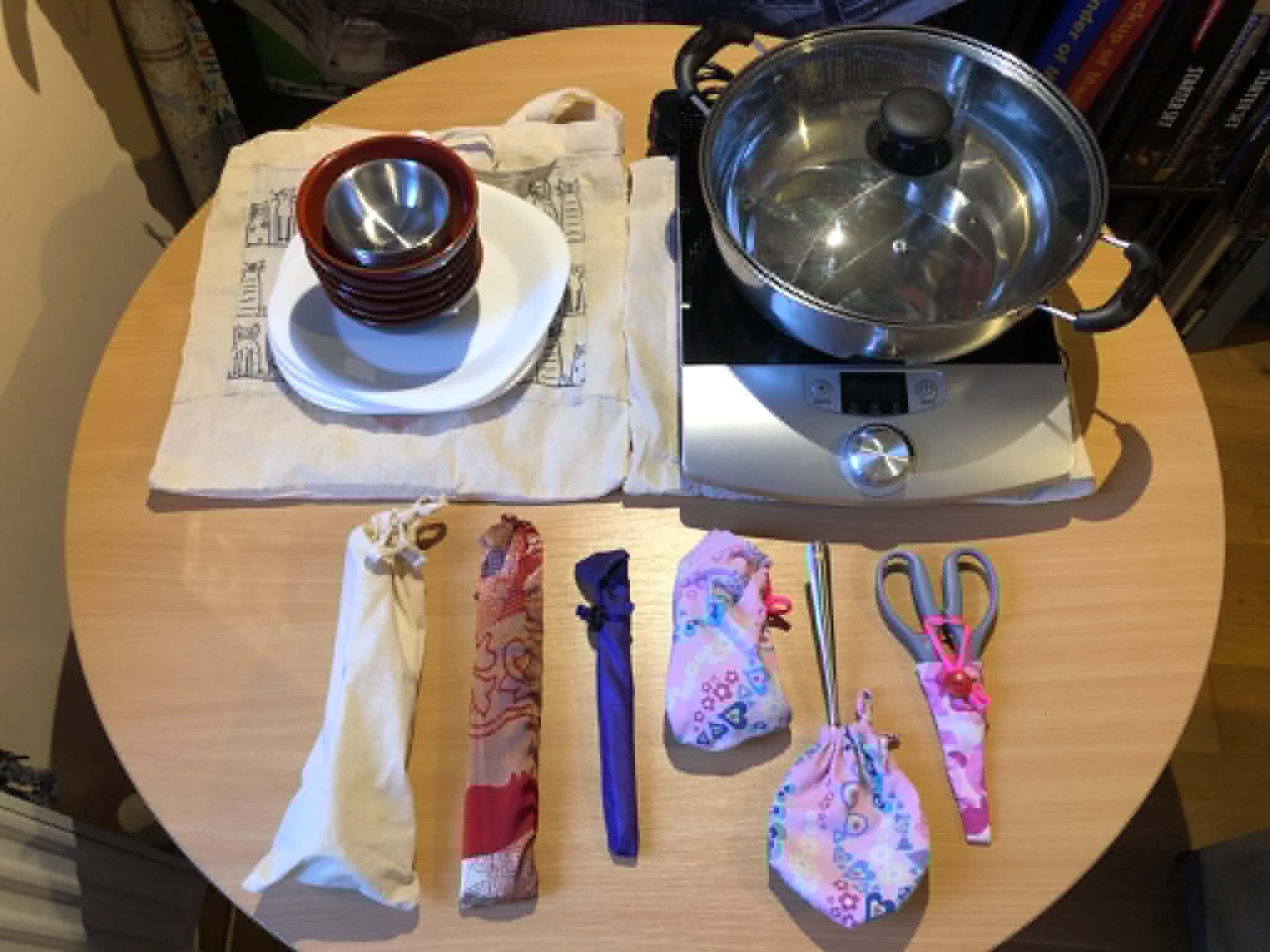 A hotpot setup including ingredient platters, bowls, pot, hob, scoops, chopsticks, spoons, skimmer, scissors and optional forks on top of their respective travel pouches.