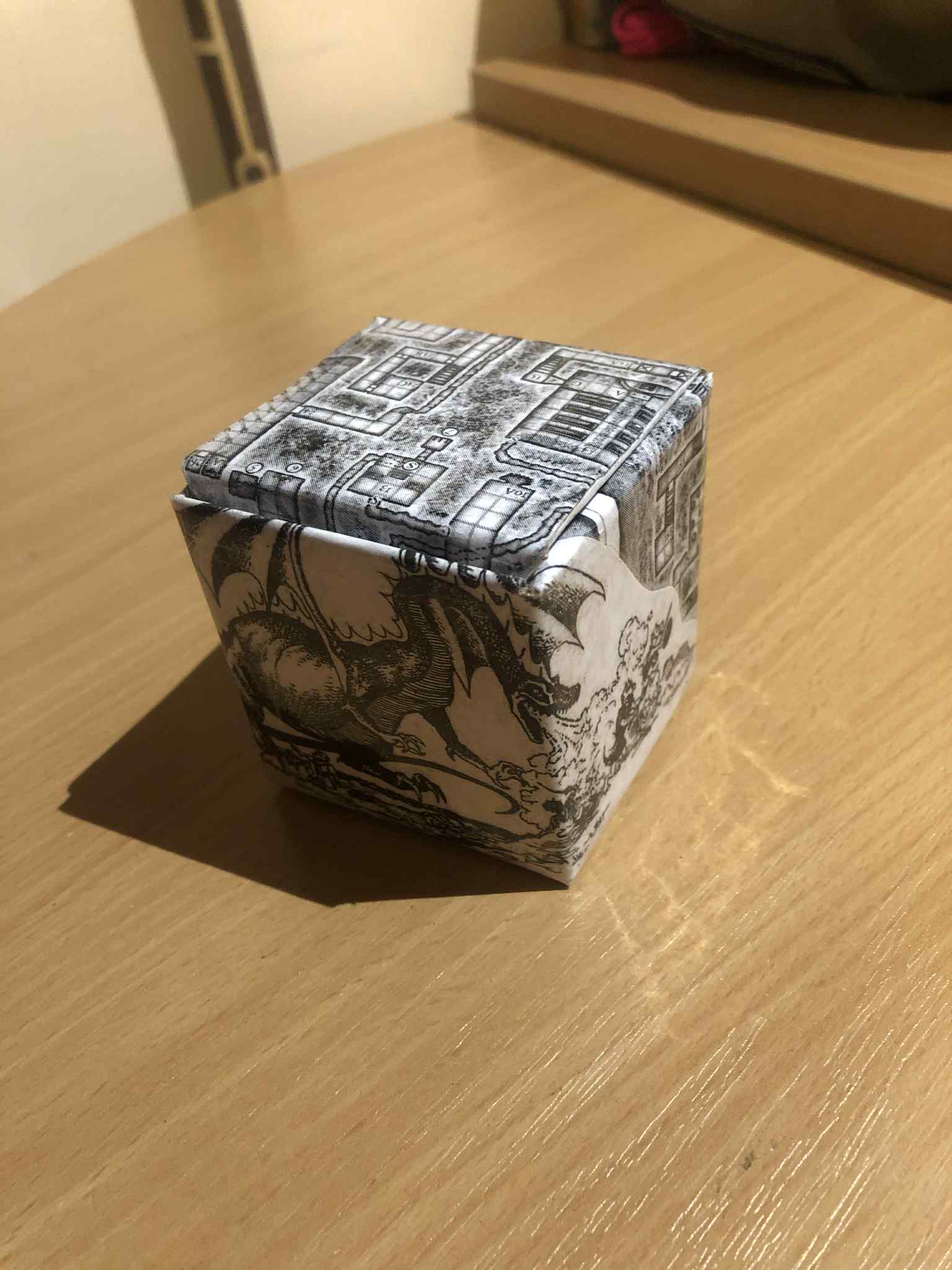 A grey looking cardboard box covered with an intricate small dungeons and dragons map over most of the box, and an illustration of a dragon breathing fire on the front.