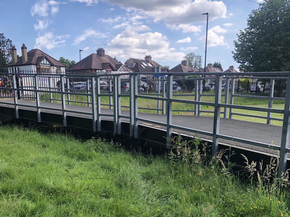 A bridge over a grassy shallow in a park, houses in the background. A space beneath the bridge beckons.