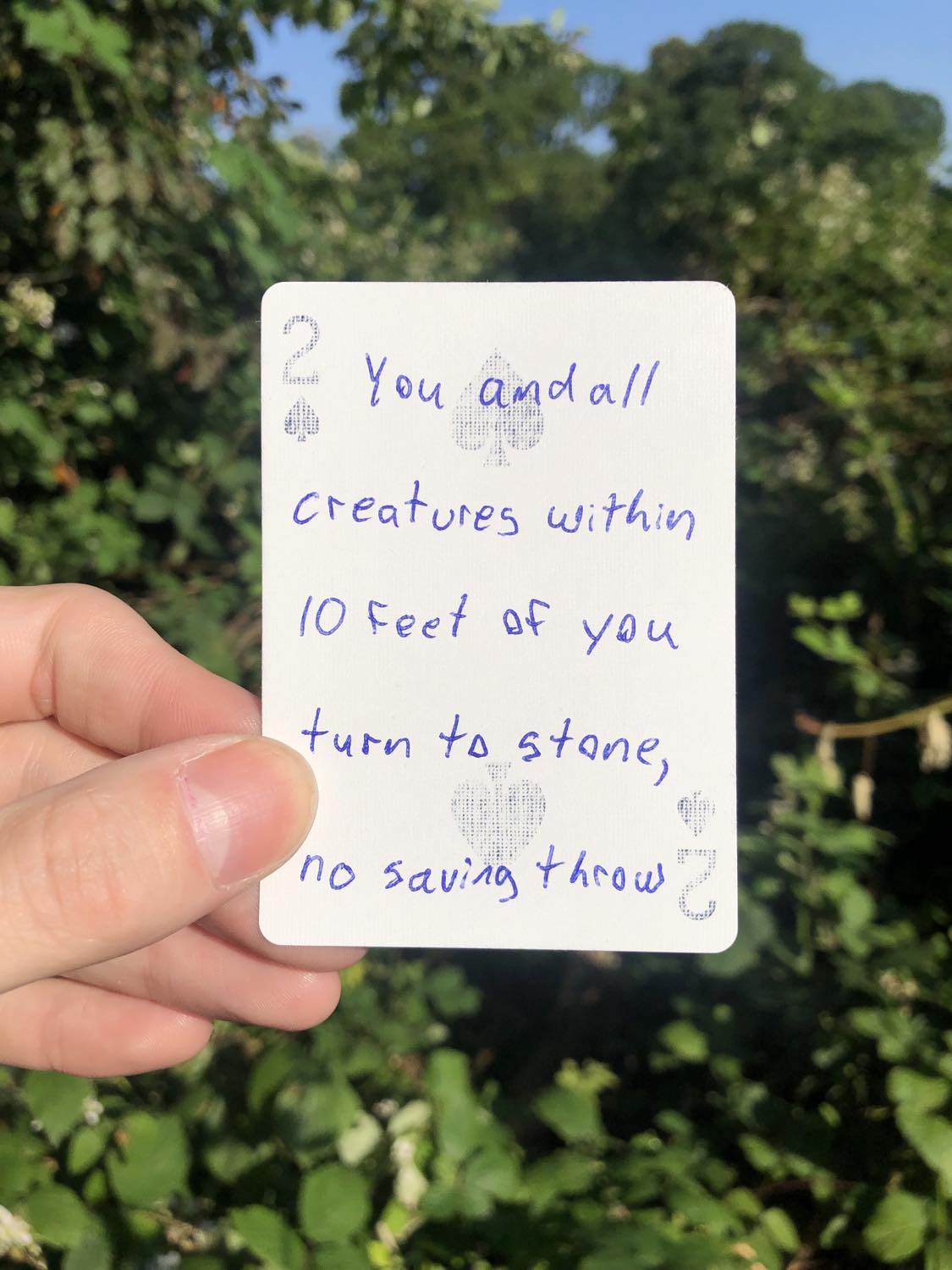 The two of spades, held with the trees and foliage behind. You and all creatures within 10 feet of you turn to stone, no saving throw.