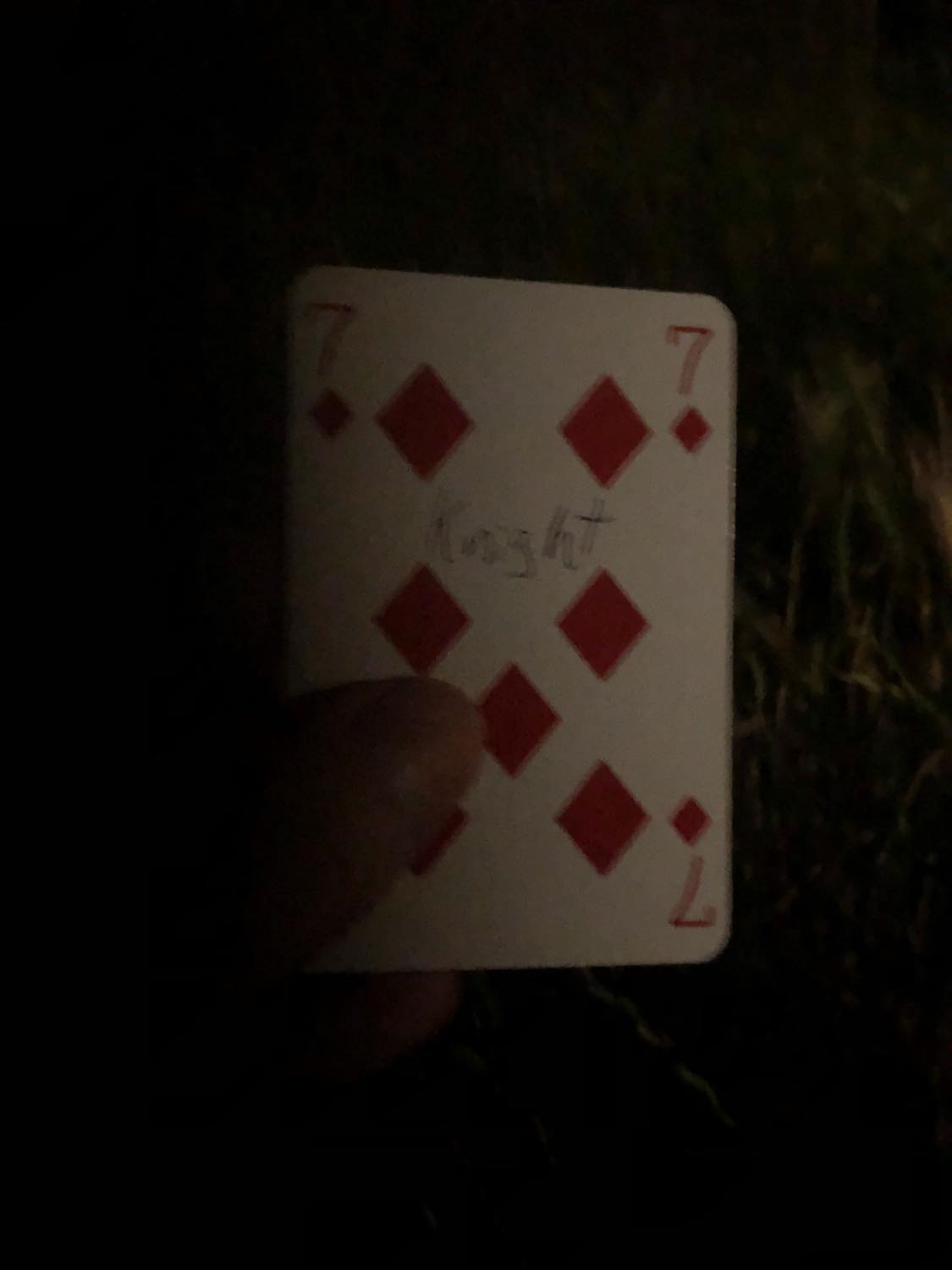 The seven of diamonds with the word Knight written on it, held in the barely lit dark.